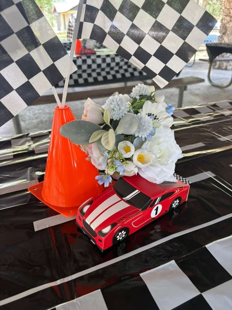 Centro de mesa temático de carreras con cono de tránsito, flores y auto miniatura sobre mantel a cuadros
