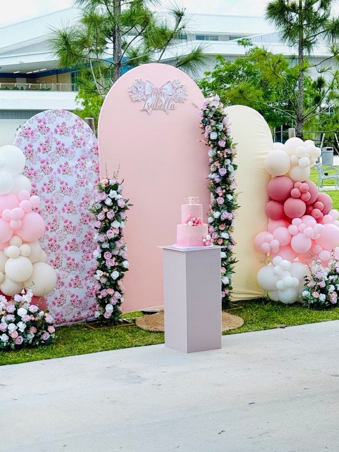 Three-arch backdrop with pink and floral panels, balloon columns, and a pink ombré cake on a white plinth